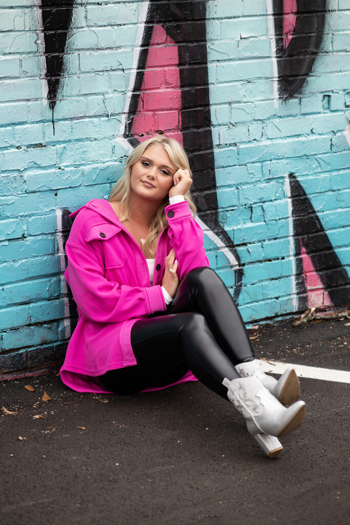 high school senior photoshoot with girl sitting agains a colorful street art wall with pink top on