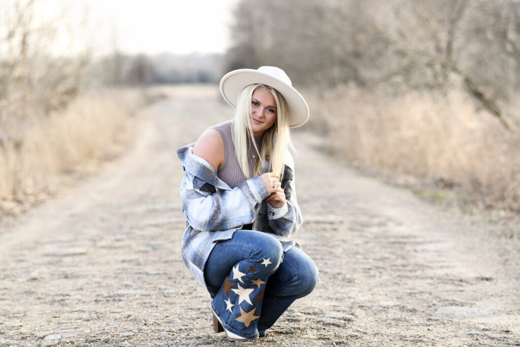 high school senior girl in middle of nature outdoor pathway crouching with rustic outfit and hat senior photo session