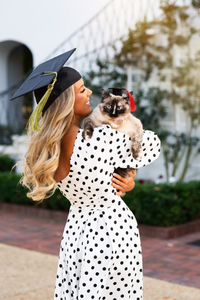 University of Alabama graduate poses holding her cat in a graduation cap in front of the President's Mansion for her graduation photos with Chris Landau Photography wearing a beautiful one shoulder white dress with black polka dots on it.