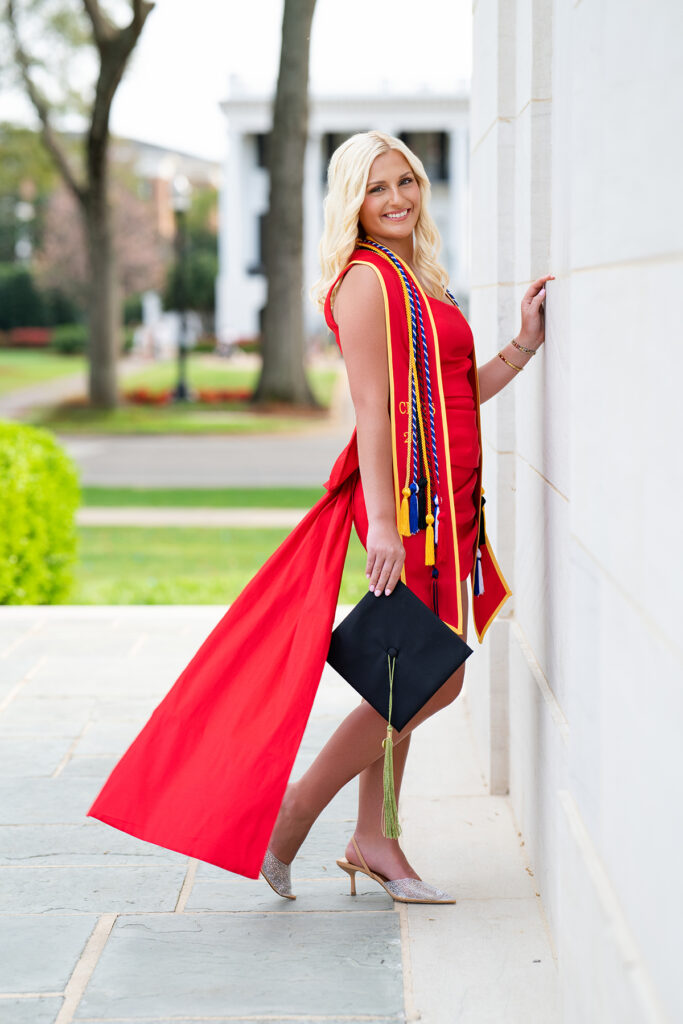 University of Alabama grad poses next to Denny Chimes for her grad photos with Chris Landau Photography wearing a gorgeous red dress with a bow on the back and long fabric tail attached to the bow