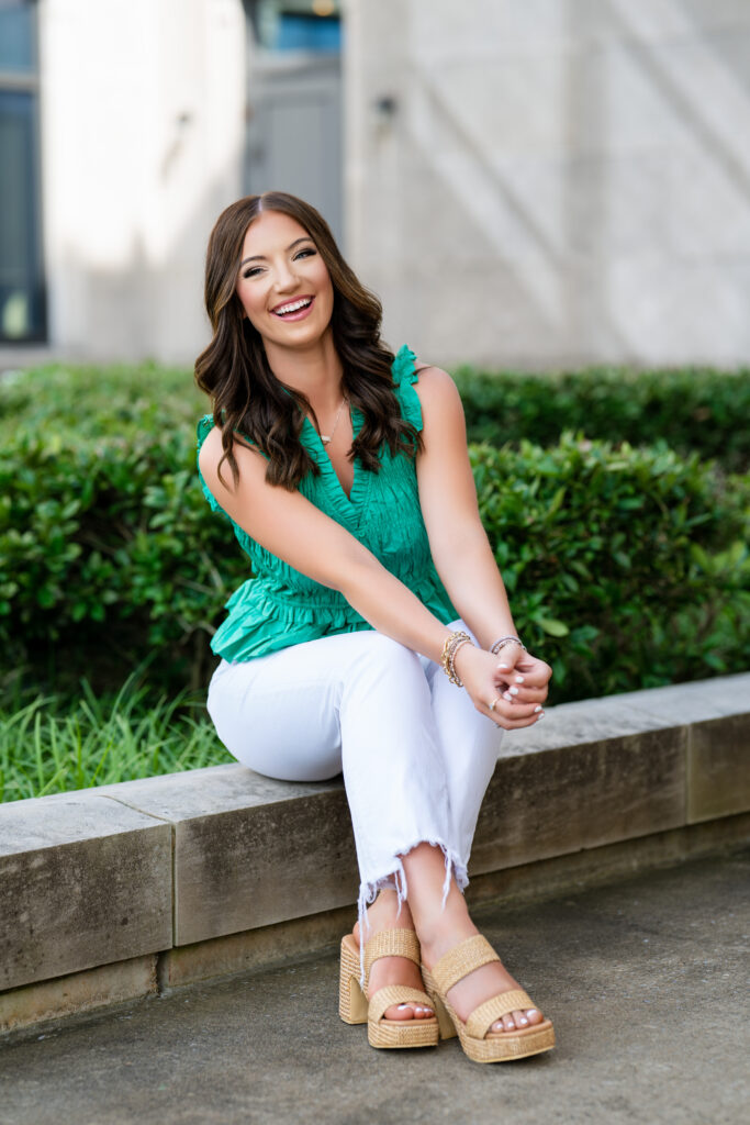 High school senior sitting on a cement wall smiling for the photo. She is wearing a green shirt and white jeans.