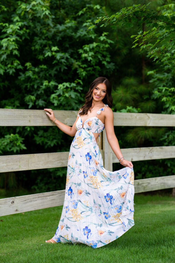 High school senior poses for her senior pictures in Tuscaloosa Alabama during her session with Chris Landau Photography. She is wearing a floral maxi-dress in a field while holding onto a fence.
