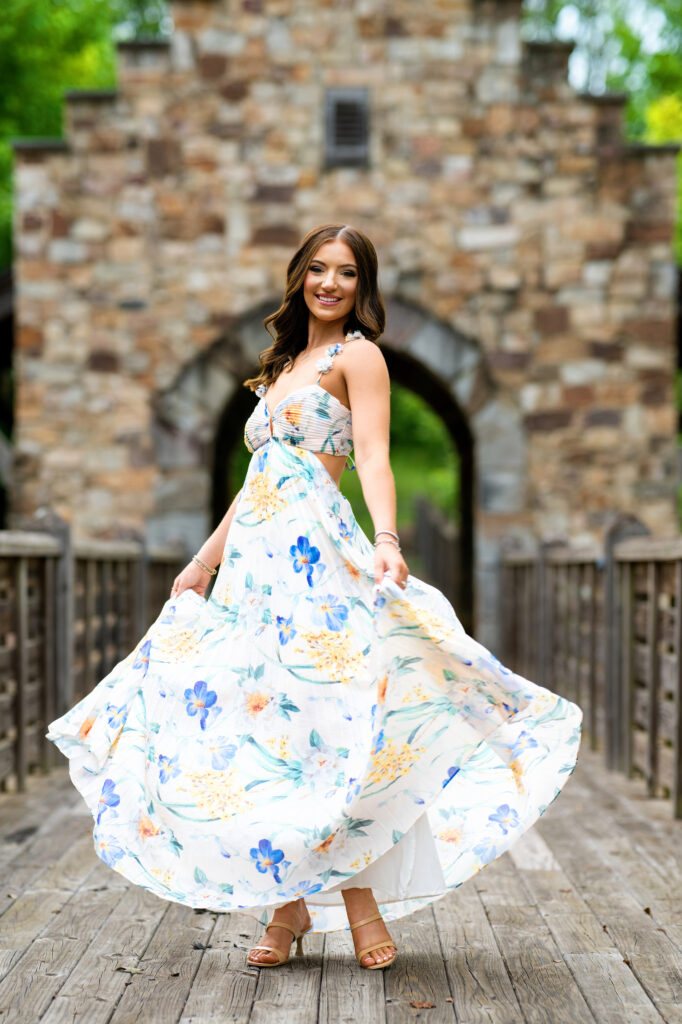 High school senior poses for her senior pictures in Tuscaloosa Alabama during her session with Chris Landau Photography. She is wearing a floral maxi-dress and twirling the skirt on a bridge in the woods.