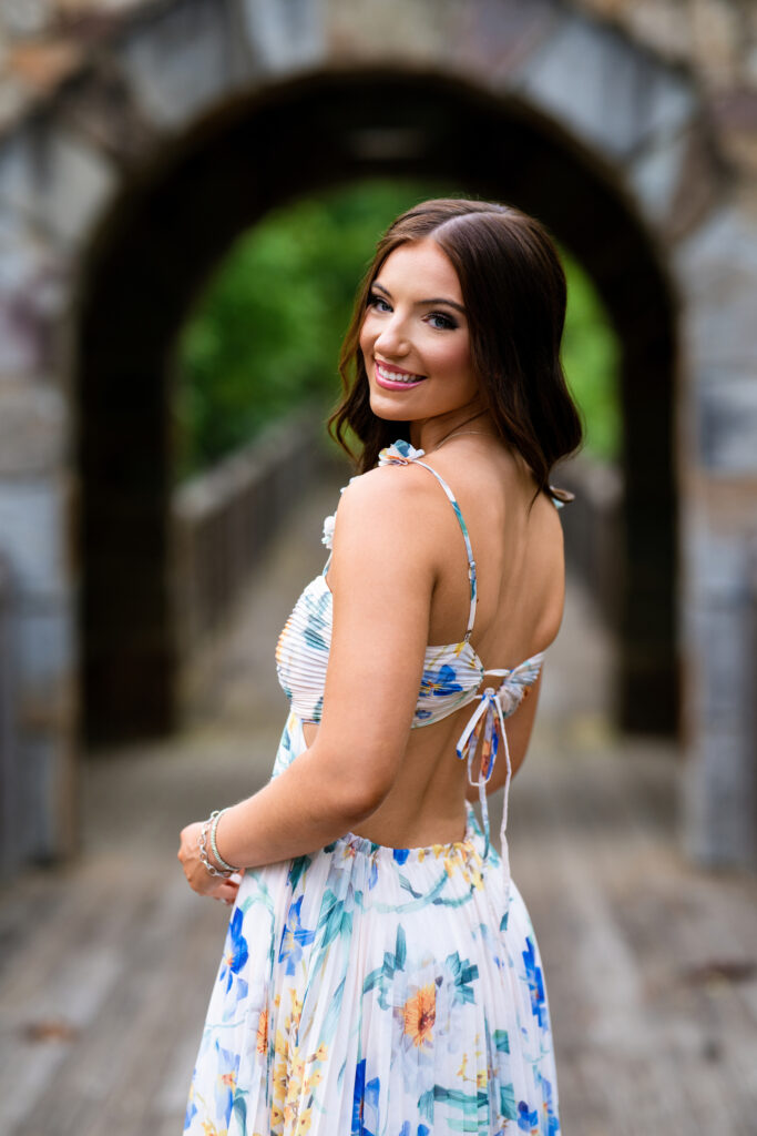 High school senior poses for her senior pictures in Tuscaloosa Alabama during her session with Chris Landau Photography. She is wearing a floral maxi-dress while standing on a bridge in nature.