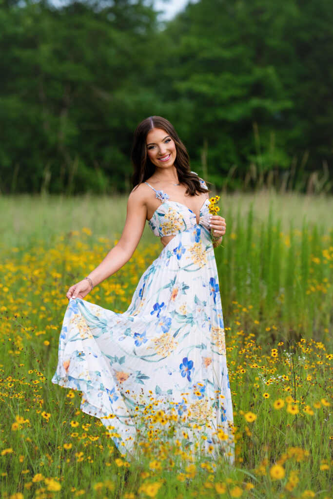 High school senior poses for her senior pictures in Tuscaloosa Alabama during her session with Chris Landau Photography. She is wearing a floral maxi-dress in a field of flowers while holding yellow flowers.