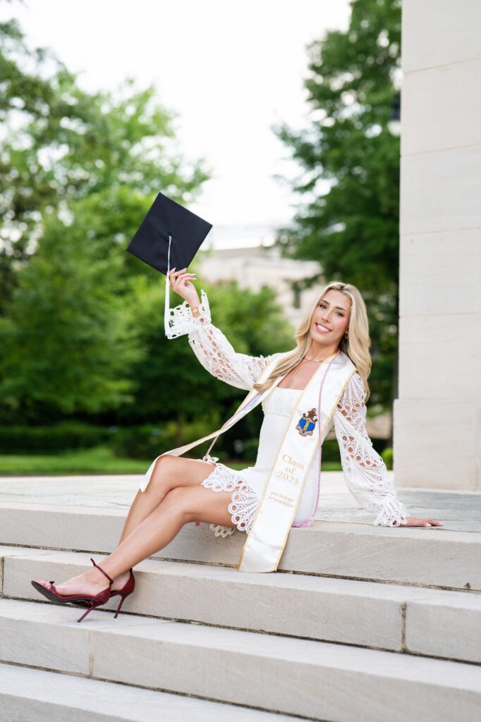 University of Alabama grad poses holding her grad cap high while sitting on steps of Denny Chimes for her photoshoot with Chris Landau Photography.