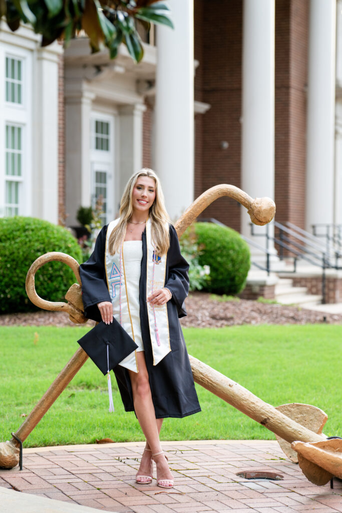 University of Alabama grad wearing her gown and Delta Gamma stole and leaning on the Delta Gamma anchor in front of her sorority house for her grad photos with Chris Landau Photography.