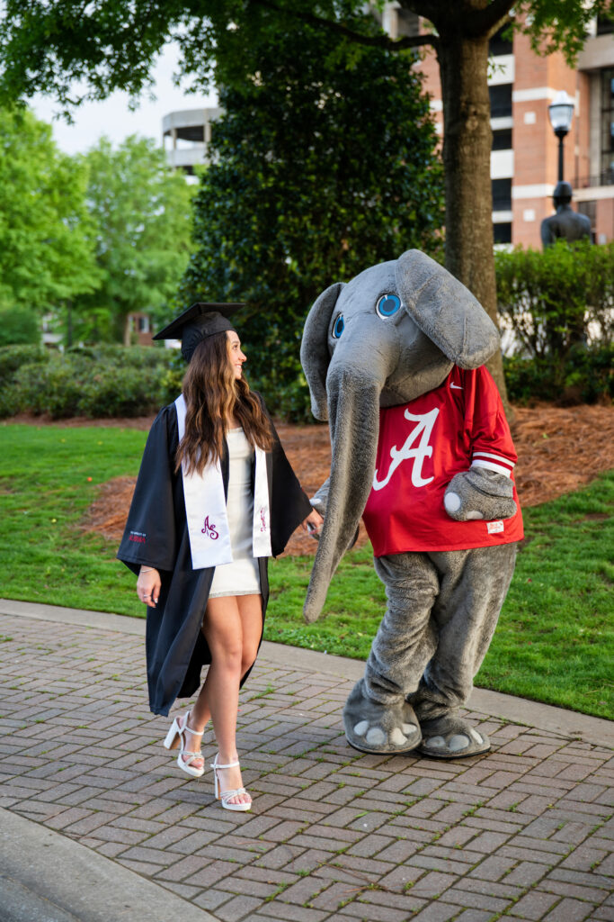 Alabama mascot Big AL walks with Alabama graduate at her graduation photoshoot with Chris landau Photography Alabama grad photographer. They are walking in front of Bryant Denny stadium on the sidewalk. Bama grad photographer