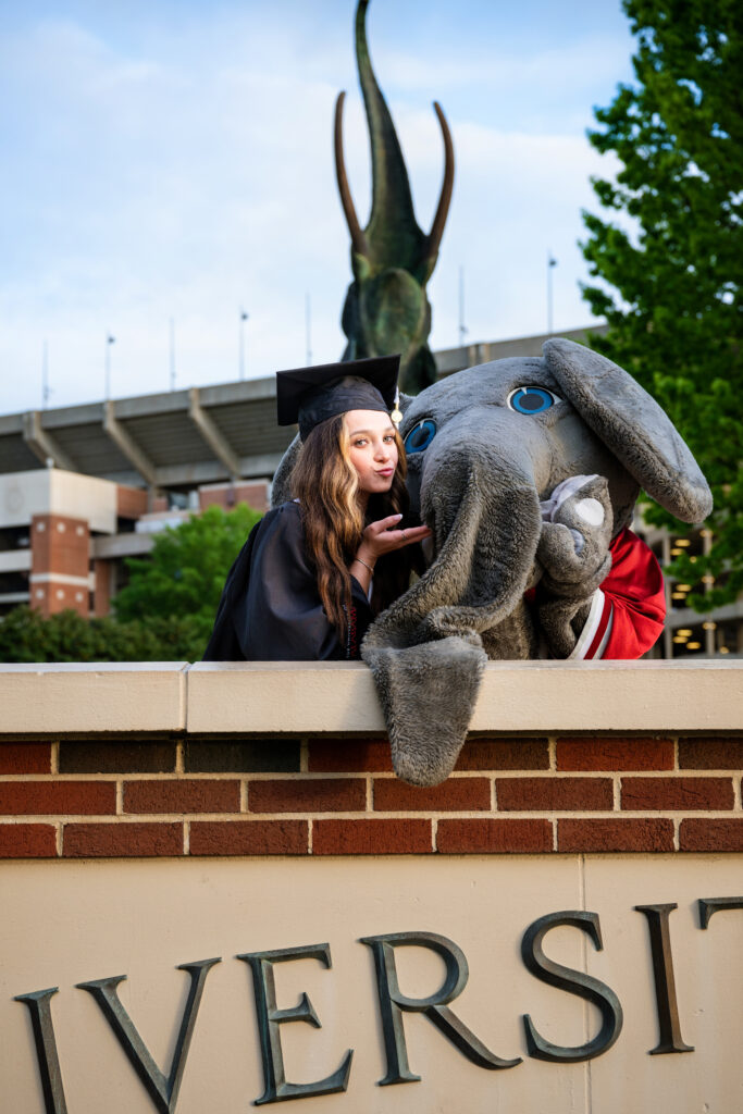 Alabama mascot Big AL poses with Alabama graduate at her graduation photoshoot with Chris landau Photography Alabama grad photographer. They are leaning on the wall in front of Tusk statue and she is blowing him a kiss. Alabama Grad photographer.