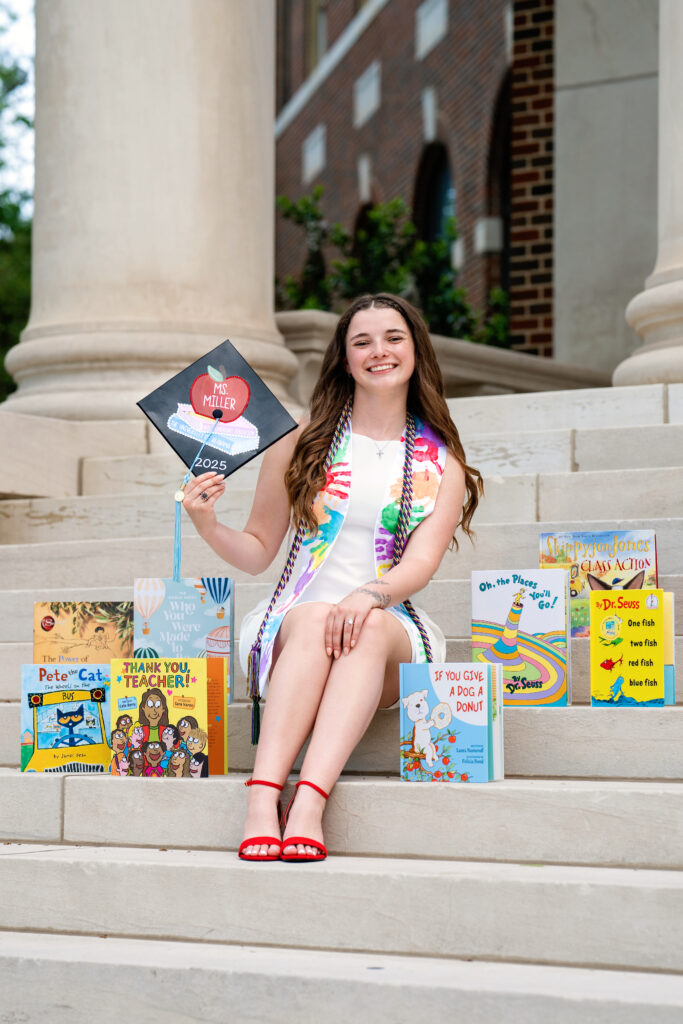 University of Alabama Elementary Education graduate sits on the steps of an academic building holding her grad cap in her hand and is surrounded by elementary school reading books.