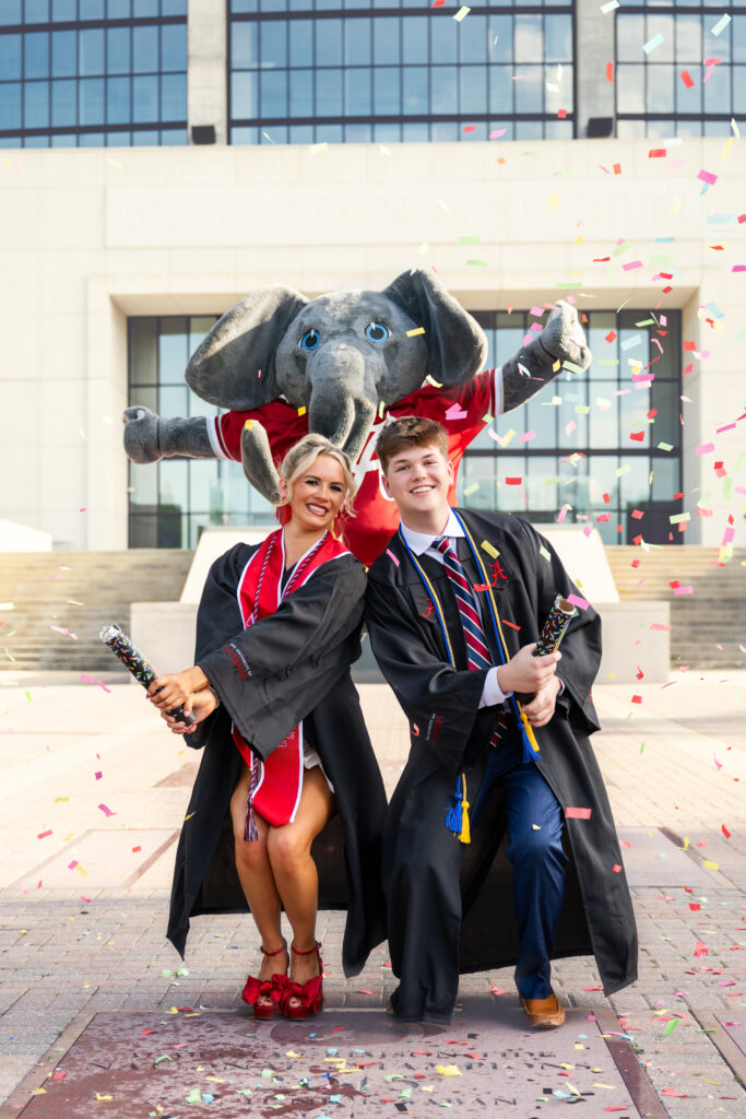 University of Alabama grad and her boyfriend use confetti while Big Al jumps behind them in front of Bryant Denny Stadium for her graduation photos with Chris Landau Photography while wearing her graduation gown