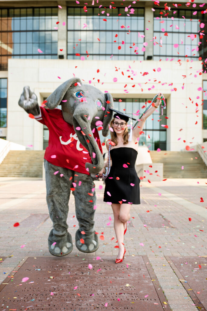 University of Alabama grad poses with Big Al the college mascot in front of Bryant Denny Stadium for her grad photos with Chris Landau Photography. Confetti is falling in front of them on the walk of champions.