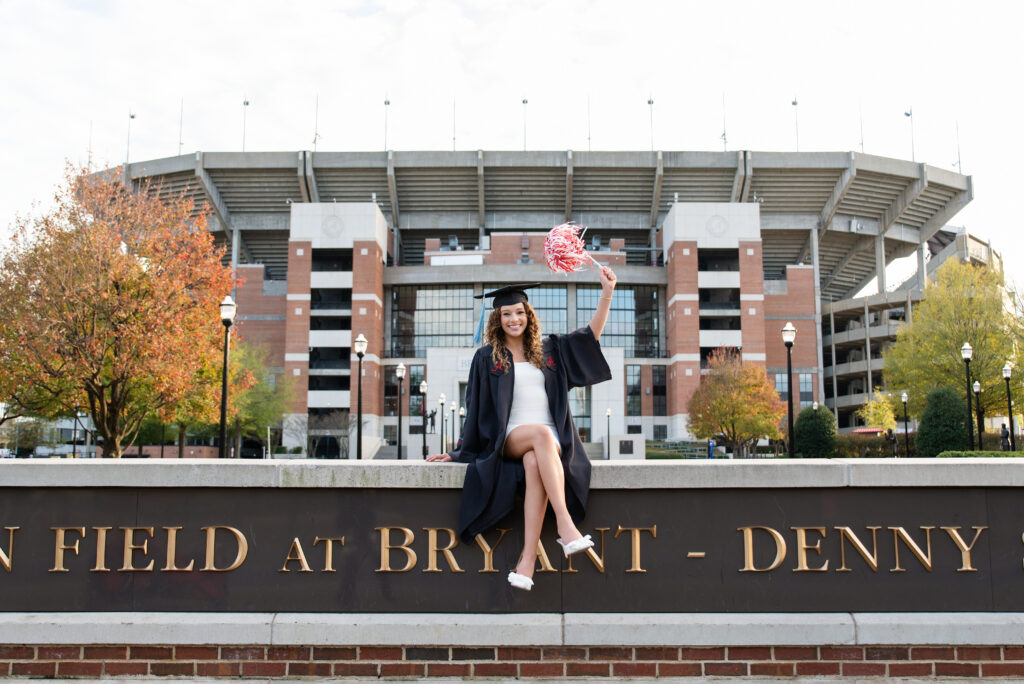 Bama grad sits on wall at Bryant Denny stadium waving a red and white shaker