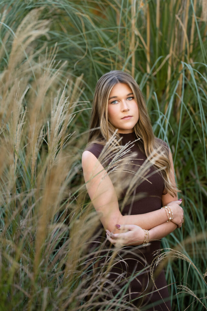 High School senior poses in tall grasses at her senior session with Chris Landau. She has a serious look with her arms crossed. 