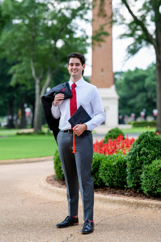 Bama grad guy poses with his gown over his shoulder and holding his cap. Denny Chimes is in the background.