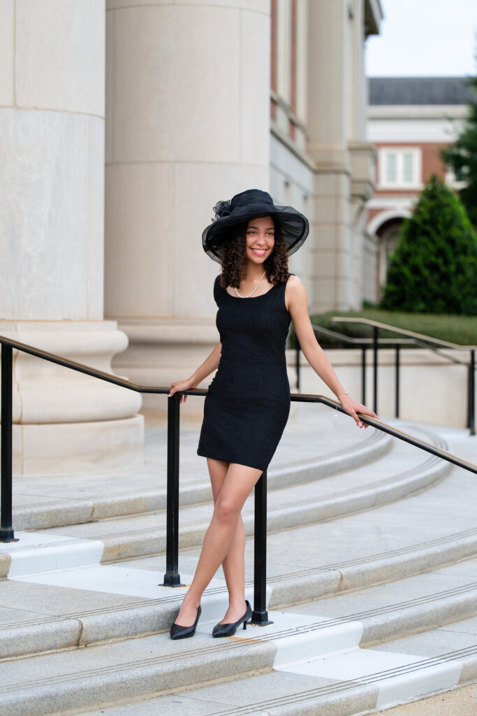 Bama grad poses at Shelby Hall wearing black dress and black hat. She is member of the honor society thirty one at Alabama